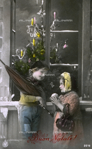 AVQ-A-002062-0060 - Postcard with a Christmas theme. Two children in modest winter clothes are posing with a window in the background through which a tree adorned with candles can be seen - Date of photography: 1914 ca. - Alinari Archives, Florence