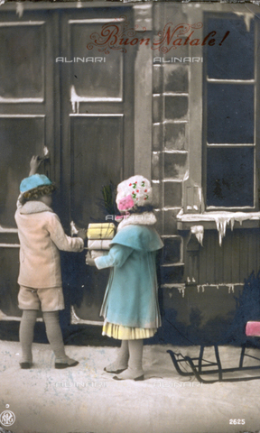 AVQ-A-002062-0076 - Christmas postcard. Two children in winter clothing are shown from behind. They are posing in the studio pretending to knock on a door which faces onto the snow-covered sidewalk. On the right there is a sled - Date of photography: 1910 ca. - Alinari Archives, Florence
