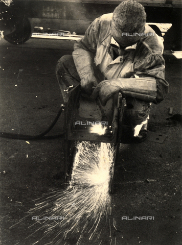 BBA-F-004775-0000 - "Oxy-hydrogen flame". Portrait of a worker using the oxy-hydrogen flame from which sparks and an intense light radiate. He is crouching before a metal object to be welded and is wearing work clothes with large gloves and protective goggles - Date of photography: 1930-1940 ca. - Alinari Archives, Florence
