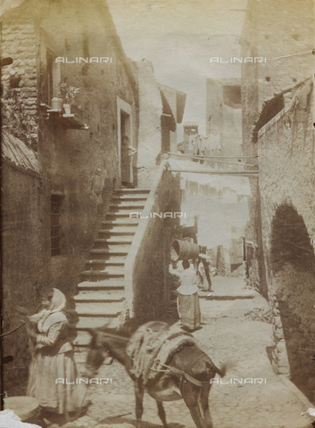 FBQ-F-001209-0000 - A small courtyard surrounded by old houses in Tivoli. In the foreground, a donkey and a woman in folk dress. In the background, a woman seen from the back is carrying a wooden container on her head - Date of photography: 1882 -1887 ca. - Alinari Archives, Florence