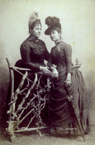 FBQ-F-002312-0000 - Full-length portrait of two ladies in elegant clothes, separated by a wooden fence. The woman in the foreground is wearing a plumed hat, with an umbrella in her left hand - Date of photography: 1887 - 1888 - Alinari Archives, Florence