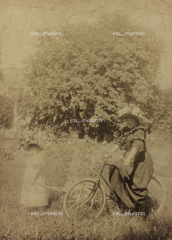 FBQ-F-004237-0000 - Lady in summer dress shown in a field, seated on a bicycle. Next to her is a little girl pulling a small wagon - Date of photography: 1890 -1900 ca. - Alinari Archives, Florence