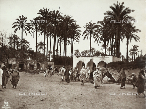 GLQ-F-002477-0000 - A view of Suk el Giuma: people in traditional clothing walk down the street, passing the low buildings. In the background: a stand of palm trees. The image dates back to the Italian conquest of Libya, just after the Italian - Turkish war of 1911-1912 - Date of photography: 1911-1912 - Alinari Archives, Florence