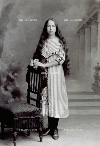 MFC-F-001161-0000 - Portrait of a girl, standing and leaning on a chair. Her long hair falls on her shoulders and she is holding a bunch of lily of the valleys - Date of photography: 1895-1910 ca. - Alinari Archives, Florence