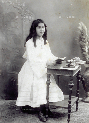 MFC-F-001260-0000 - Full-length portrait of young girl sitting at a table, with a book in her hands. Some stationery can be seen on the table such as letters and a paperweight - Date of photography: 1900-1920 ca. - Alinari Archives, Florence