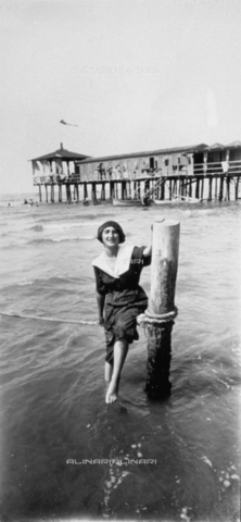 MFC-F-001735-0000 - Young woman in beachwear, sitting on a rope tied to a pole which sticks out of the water, a few steps from the shore, on the beach of Viareggio. In the background the pier, with a few cabins - Date of photography: 1900-1910 ca. - Alinari Archives, Florence