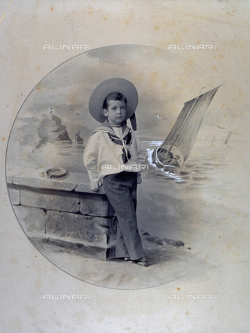 MFC-F-002078-0000 - Full-length portrait of a child, leaning against a low wall. The painted background shows a marine landscape with rocks, lighthouse and sailboat in the foreground - Date of photography: 1900-1910 ca. - Alinari Archives, Florence