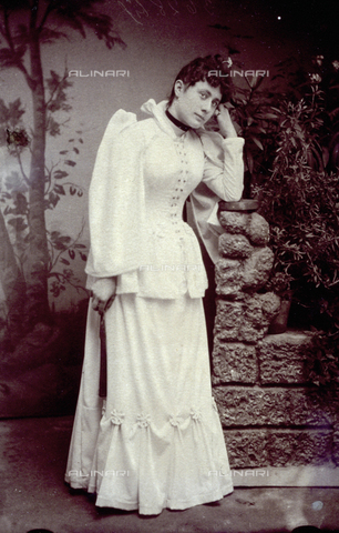 MFC-F-004040-0000 - Full-length portrait of a young woman in a white dress. She is shown against a painted backdrop and is leaning against a fake low wall with plants on it - Date of photography: 1880-1885 ca. - Alinari Archives, Florence