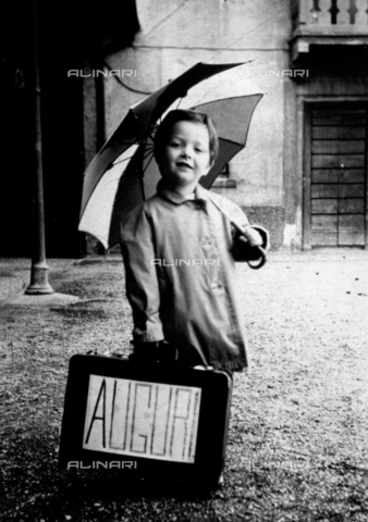 PDC-F-004209-0000 - Ritratto a figura intera di bambino dal volto sorridente. Il piccolo indossa un impermeabile e tiene un ombrello aperto; con la mano destra sostiene una valigia con la scritta 'Auguri' - Data dello scatto: 1949-1951 - Archivi Alinari, Firenze